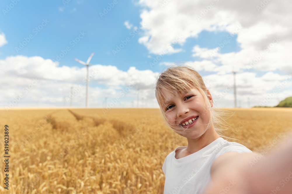 Portrait of little cute beautiful blond caucasian girl enjoy having fun taking selfie photo phone camera walking at wheat field against wind mill turbine farm. Children blogging and video streaming Portrait of little cute beautiful blond caucasian girl enjoy having fun taking selfie photo phone camera walking at wheat field against wind mill turbine farm. Children blogging and video streaming