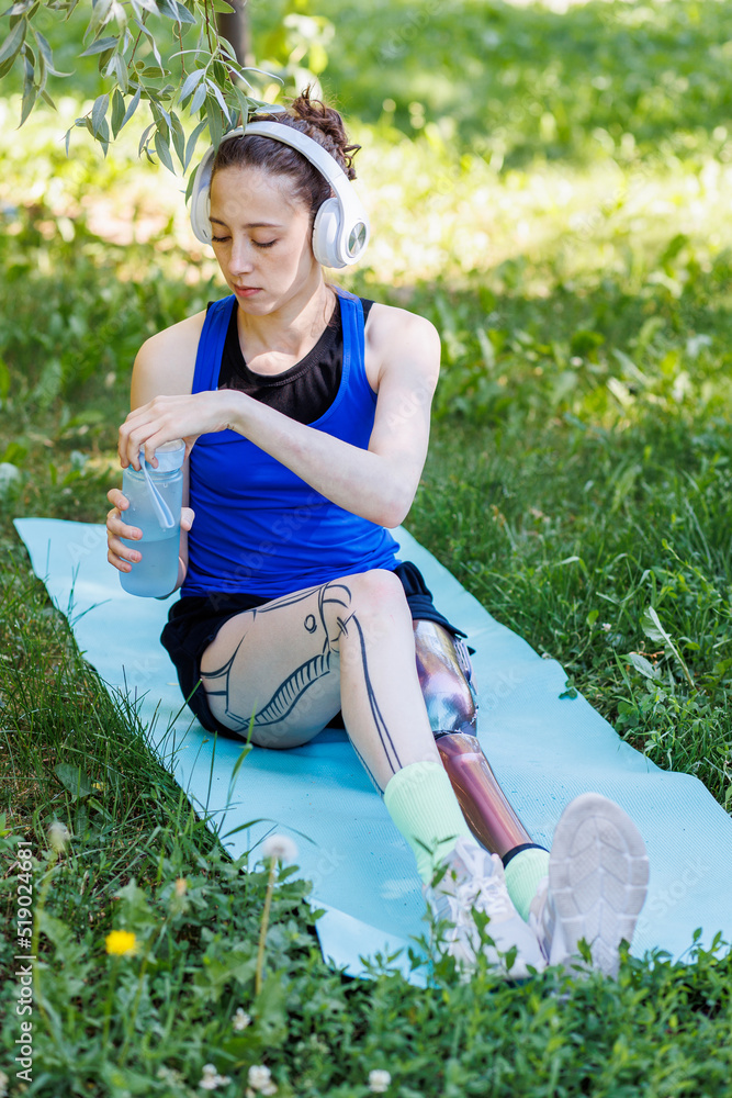 Young beautiful athletic woman with bionic prosthetic leg doing sport  exercises in the park outdoors in summer. Concept of active lifestyle of  disabled people. Listening to music, drinking water Stock Photo