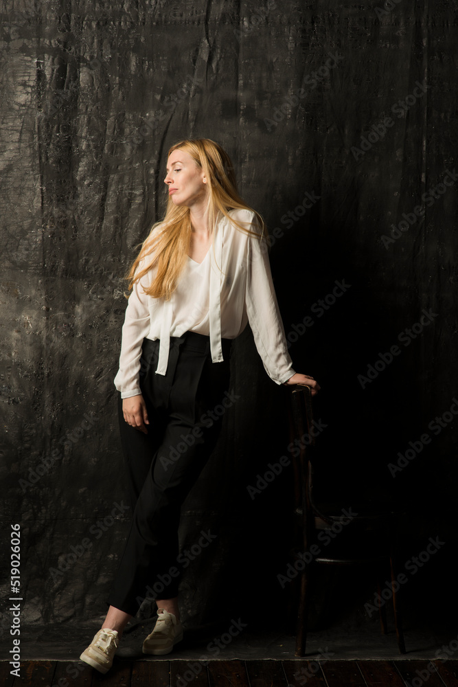 portrait of a beautiful blonde woman with long hair in a white blouse and black trousers on a gray isolated background.a charming girl posing on a dark background. portrait of a beautiful blonde woman with long hair in a white blouse and black trousers on a gray isolated background.a charming girl posing on a dark background.