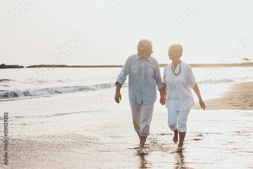 Couple of old mature people walking on the sand together and having fun on the sand of the beach enjoying and living the moment. Two cute seniors in love having fun. Barefoot walking on the water.