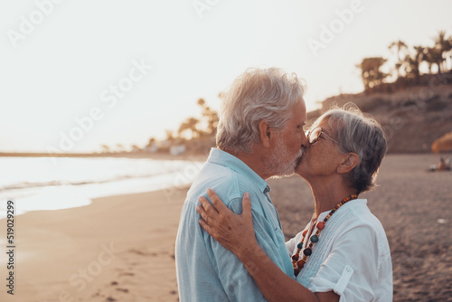 Close up of two cute and happy seniors having fun and enjoying together a sunset day at the beach. Mature couple in love kissing together with the sunset at the background..