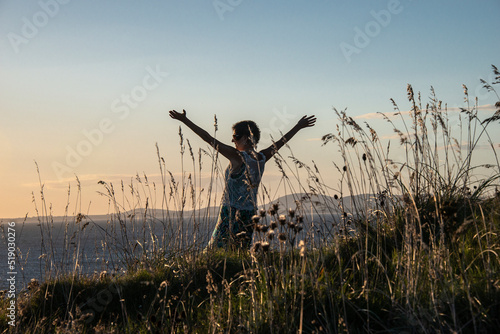 Mature woman in Punta Ballena, standing among weeds with her arms outstretched in front of the sea at sunset, with the Piriápolis hills in the background, in the department of Maldonado, Uruguay. photo