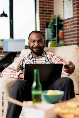 African american man working on laptop and switching tv channels with remote control, browsing internet and finding film to watch. Eating takeout delivery fast food meal at home.