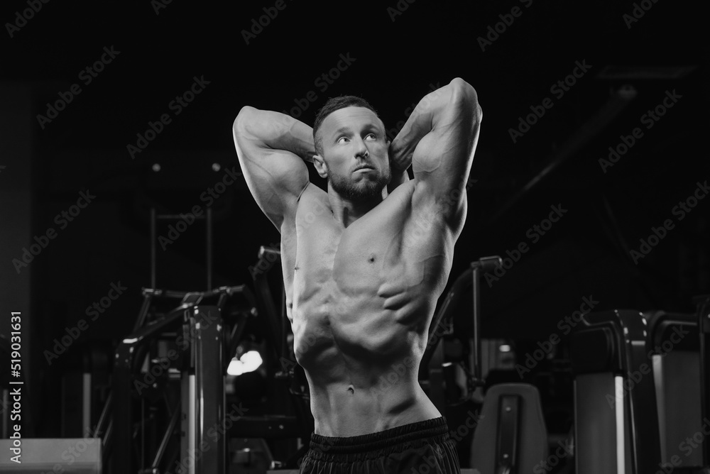 A bodybuilder with a beard is doing a stomach vacuum pose after a workout in a gym.