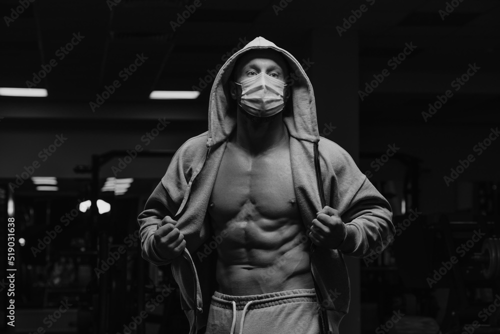 A bodybuilder in a hood and a face mask to avoid the spread of coronavirus is opening a hoodie to demonstrate his athletic physique. A sporty guy in a surgical mask is posing after a workout in a gym.