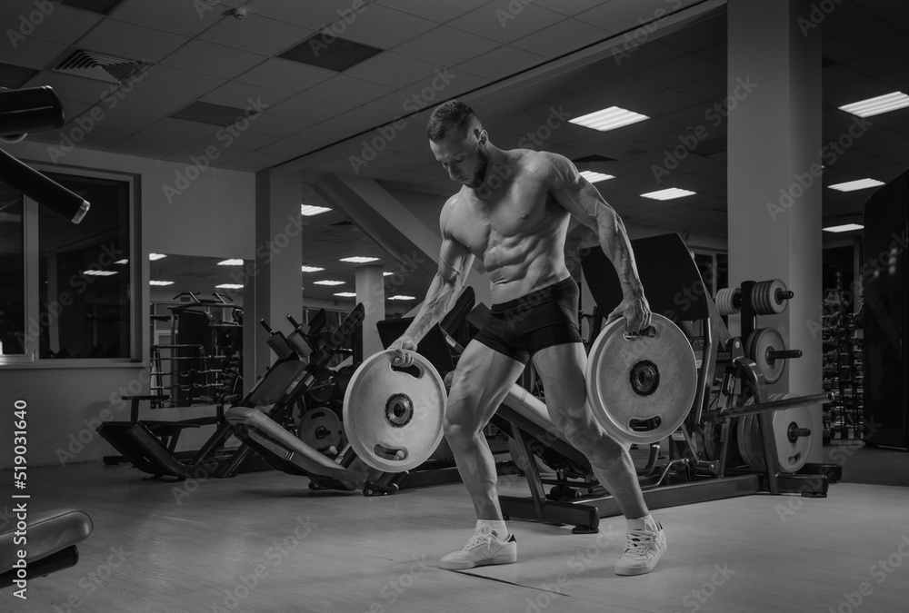 A bodybuilder in shorts is walking through the gym with two rubber plates. A shirtless sporty guy is training alone.