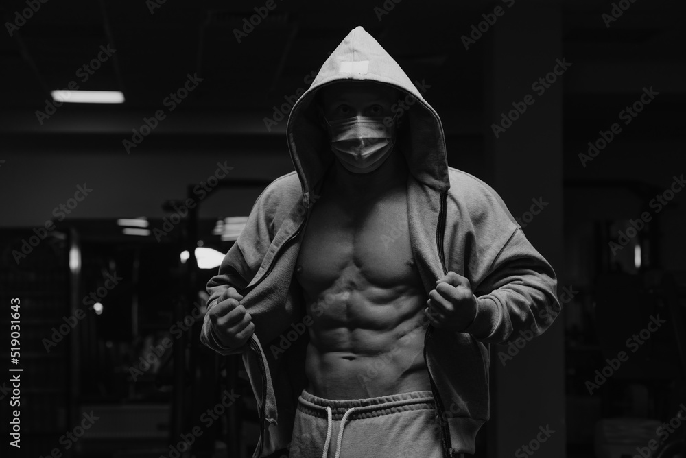 A bodybuilder in the hood and a face mask to avoid the spread of coronavirus is opening his zipped hoodie to demonstrate his abs. A sporty guy in a surgical mask is posing after a workout in a gym.