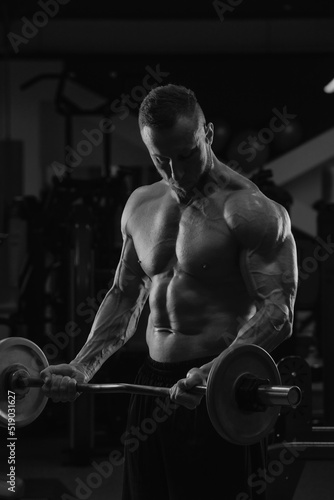 Obraz na płótnie A bodybuilder with a beard is doing bicep curls with a short barbell in a gym. A photo of a torso of a naked muscular man during a workout. w salonie