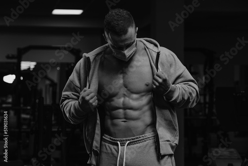 A bodybuilder in a face mask to avoid the spread of coronavirus is opening his zipped hoodie to demonstrate his athletic physique. A sporty guy in a surgical mask is posing after a workout in a gym.
