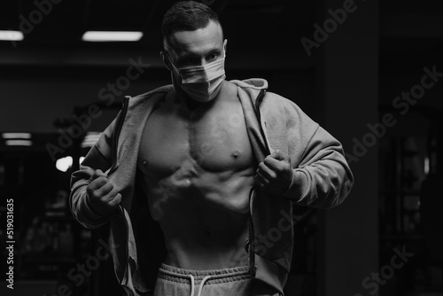 Obraz na płótnie A bodybuilder in a face mask to avoid the spread of coronavirus is opening his zipped hoodie to demonstrate his vacuum workout. A sporty guy in a surgical mask is posing after training in a gym. w salonie