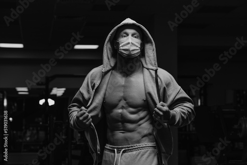 Obraz na płótnie A bodybuilder in a hood and a face mask to avoid the spread of coronavirus is opening a hoodie to demonstrate his athletic physique. A sporty guy in a surgical mask is posing after a workout in a gym. w salonie