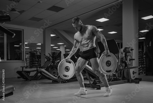 Obraz na płótnie A bodybuilder in shorts is walking through the gym with two rubber plates. A shirtless sporty guy is training alone. w salonie