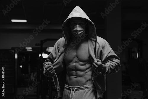 Obraz na płótnie A bodybuilder in the hood and a face mask to avoid the spread of coronavirus is opening his zipped hoodie to demonstrate his abs. A sporty guy in a surgical mask is posing after a workout in a gym. w salonie