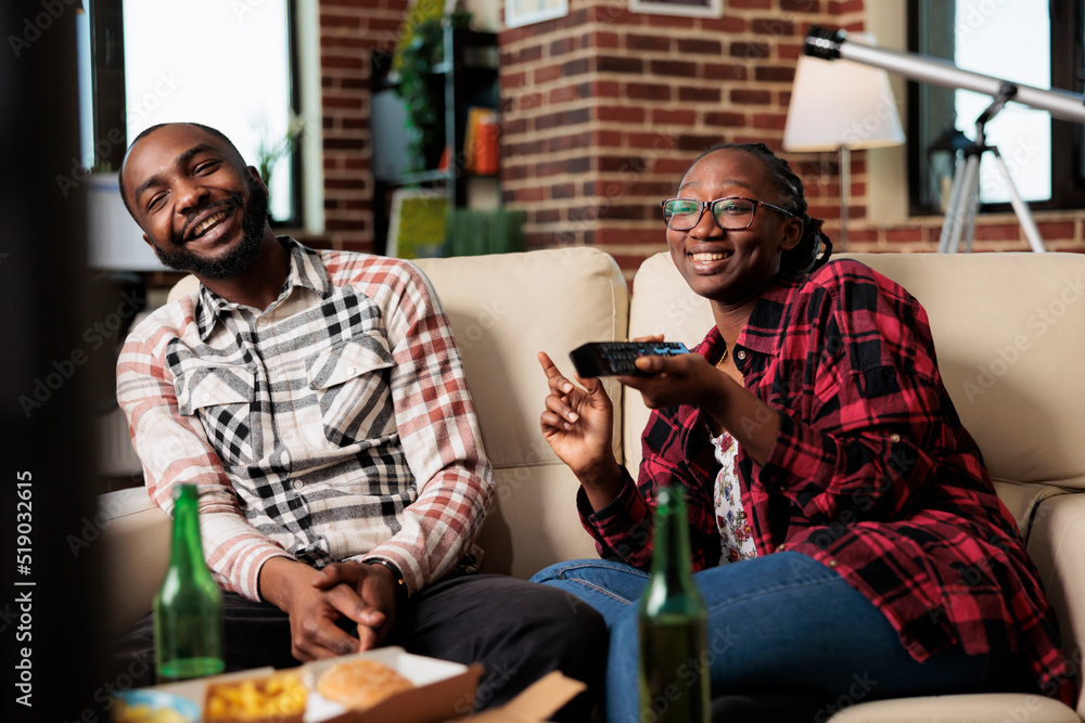 Cheerful couple laughing and watching television together, eating fast food delivery meal while girlfriend switches channels with tv remote control. Takeaway food and beer to have fun with film.