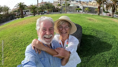 Cute couple of old and mature people having fun together at the park with the beach at the background. Two seniors jumping and looking at the camera taking a selfie together.
