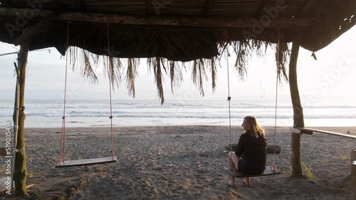 Young blonde woman enjoying a beautiful sunset while sitting on a swing set at a beach in Costa Rica. Smooth and stable push through shot photo