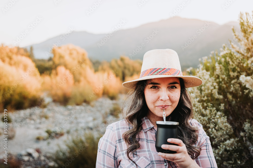 Smiling woman with a hat and wearing a pale pink blouse, drinking mate in a natural space at sunset Smiling woman with a hat and wearing a pale pink blouse, drinking mate in a natural space at sunset