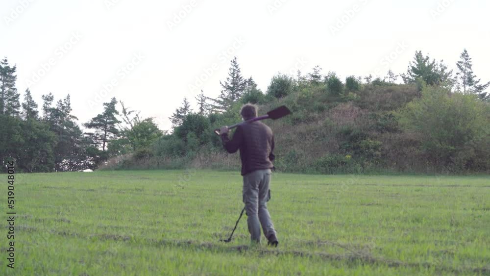 Man Walking With Shovel On His Shoulder, Detecting Metals Buried On The Ground Near Tumulus Using Metal Detector Device. wide Man Walking With Shovel On His Shoulder, Detecting Metals Buried On The Ground Near Tumulus Using Metal Detector Device. wide