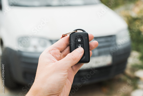 Close-up of human hand using car key remote control to unlock an alarm. Selective focus on remote control, white car in bokeh