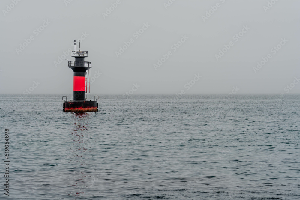 Red and black lighthouse in waters offshore on overcast morning.