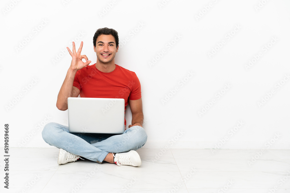Caucasian handsome man with a laptop sitting on the floor showing ok sign with fingers Caucasian handsome man with a laptop sitting on the floor showing ok sign with fingers