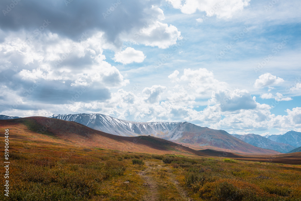 Motley autumn landscape with hills on high plateau and sunlit snowy mountain range under dramatic cloudy sky. Vivid autumn colors in mountains. Sunlight and shadows of clouds in changeable weather. Motley autumn landscape with hills on high plateau and sunlit snowy mountain range under dramatic cloudy sky. Vivid autumn colors in mountains. Sunlight and shadows of clouds in changeable weather.