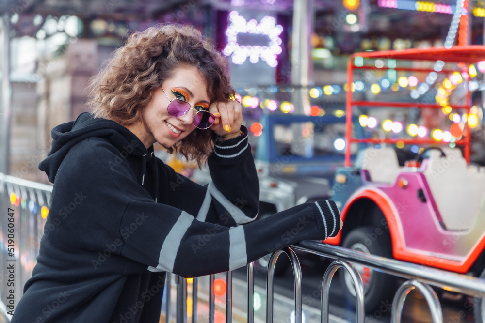 Young cool woman with curly hair and bright make-up in sunglasses posing in amusement park. Street fashion, trendy outfit Young cool woman with curly hair and bright make-up in sunglasses posing in amusement park. Street fashion, trendy outfit