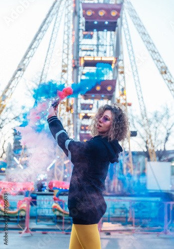 Bright cool woman with colored makeup and curly hair posing with colored smoke in the amusement park. Street fashion © splitov27