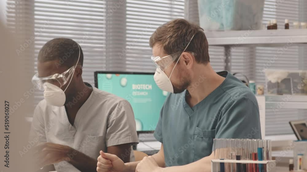 Medium slowmo of two multiethnic male biotechnologists in scrubs, respiratory masks and protective glasses conducting experiments with substances in laboratory Medium slowmo of two multiethnic male biotechnologists in scrubs, respiratory masks and protective glasses conducting experiments with substances in laboratory