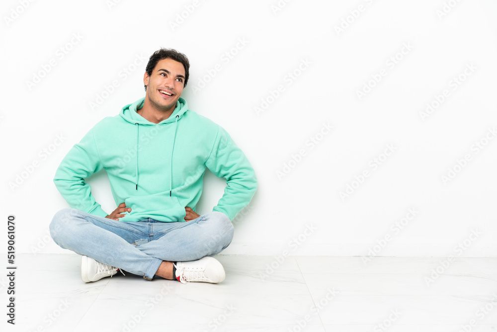 Caucasian handsome man sitting on the floor posing with arms at hip and smiling