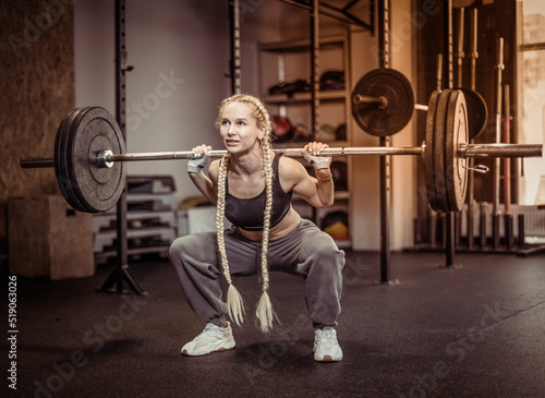 Young fit woman doing squats with heavy barbell on her shoulders in modern gym. Functional, training muscles legs and buttocks