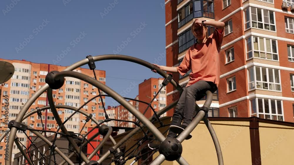 A teenage girl on a playground in the yard, she sits on top of a metal structure with ropes. A girl climbs on ropes and pipes on a modern sports ground in the courtyard of the house. A teenage girl on a playground in the yard, she sits on top of a metal structure with ropes. A girl climbs on ropes and pipes on a modern sports ground in the courtyard of the house.