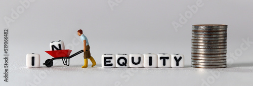 White cube with the word 'EQUITY'. A miniature man carrying a white cube with an 'IN' on it in a cart. The concept of inequity and equity.
