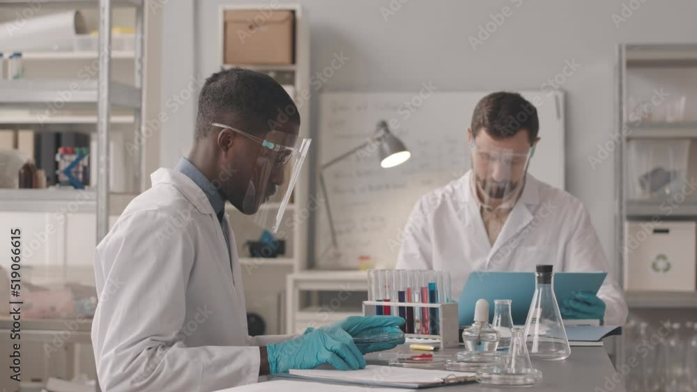 Medium slowmo of two diverse male chemists wearing lab coats and protective face shields sitting at table with glass equipment on it and doing research with substances in modern laboratory Medium slowmo of two diverse male chemists wearing lab coats and protective face shields sitting at table with glass equipment on it and doing research with substances in modern laboratory