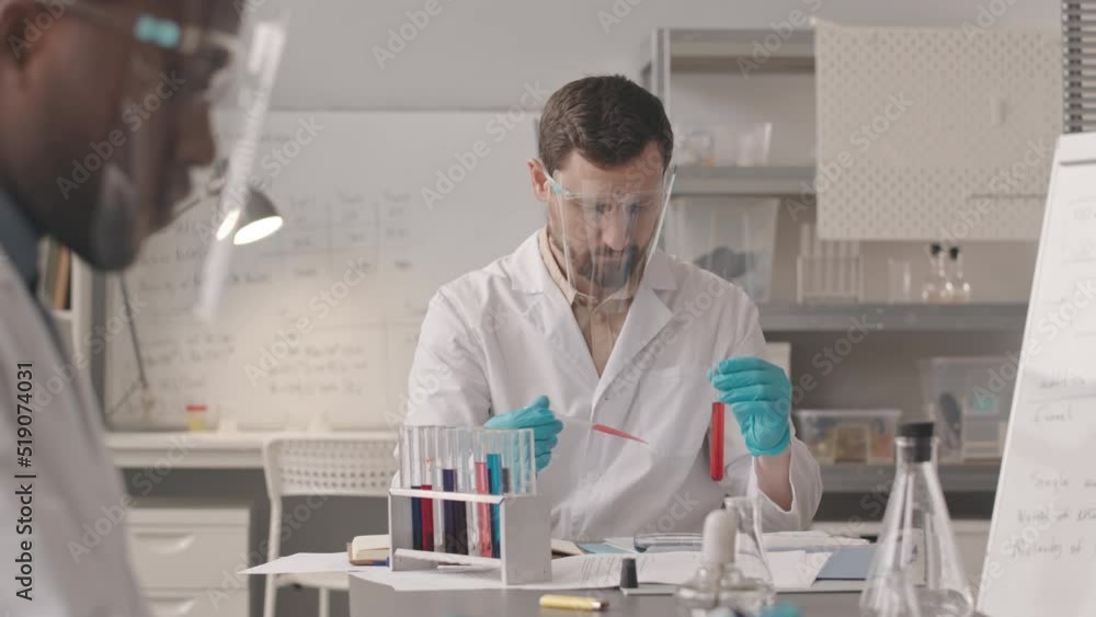 Medium slowmo of Caucasian male chemist in workwear dropping red liquid substance from test tube to petri dish with pipette doing research, working in scientific lab with African American colleague Medium slowmo of Caucasian male chemist in workwear dropping red liquid substance from test tube to petri dish with pipette doing research, working in scientific lab with African American colleague