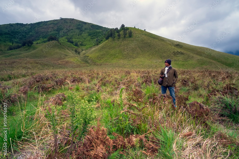 Photo of an asian man standing in the middle of meadow in mount bromo on a cloudy morning
