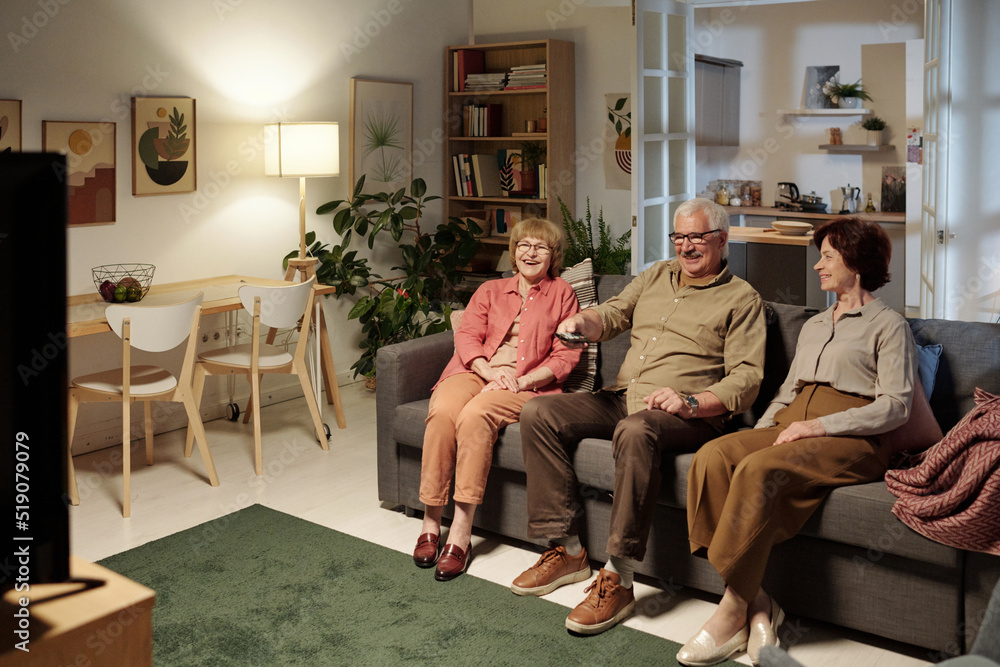 Three aged friends in casualwear sitting on sofa in front of tv set while man with remote control in hand choosing channel Three aged friends in casualwear sitting on sofa in front of tv set while man with remote control in hand choosing channel