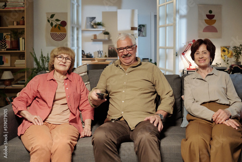 Group of happy senior friends in casualwear sitting on comfortable couch in living room and watching tv while relaxing at home together