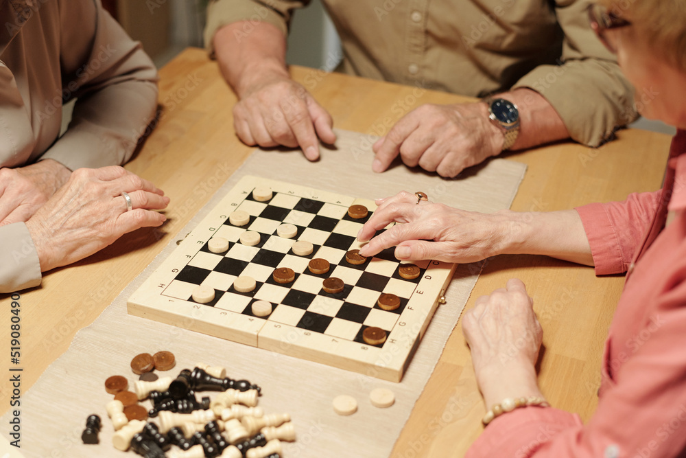 Hands of three senior friendly people gathered by table with checkers, chess figures and chessboard to play leisure games Hands of three senior friendly people gathered by table with checkers, chess figures and chessboard to play leisure games