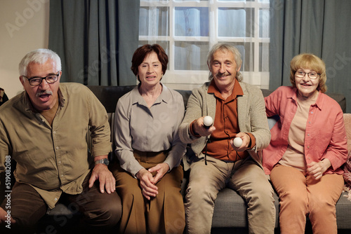 Group of cheerful mature friends sitting on sofa in living room in front of tv set while playing video game and commenting their actions