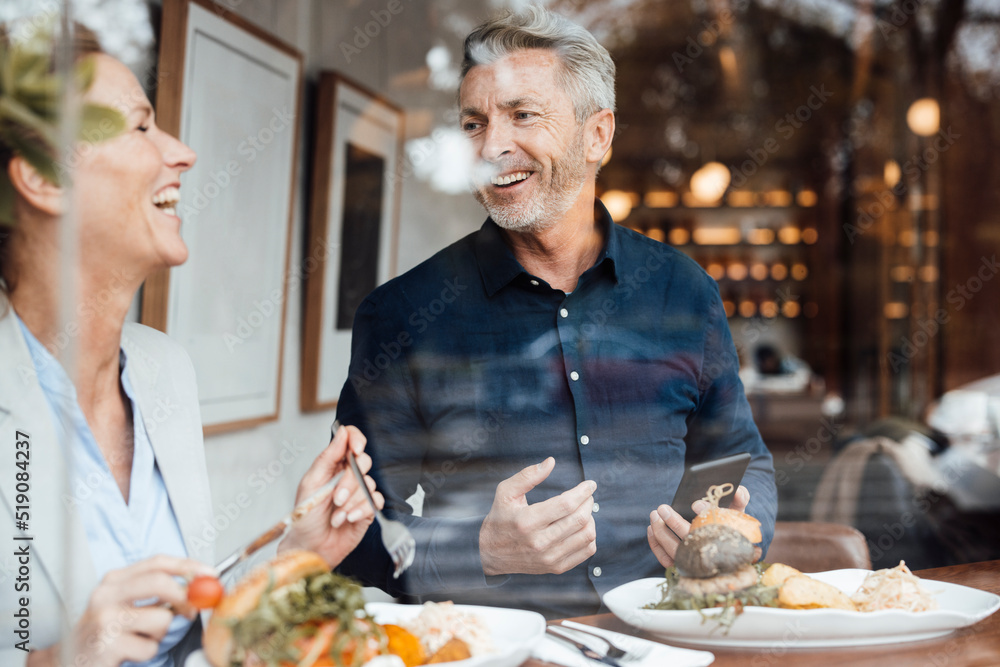 Happy businesswoman with businessman holding mobile phone having lunch in cafe seen through glass Happy businesswoman with businessman holding mobile phone having lunch in cafe seen through glass