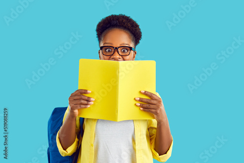Happy cheerful smart Afro American high school, college or university student girl in eyeglasses isolated on blue background hides half of her face behind book or diary with notes and looks at camera