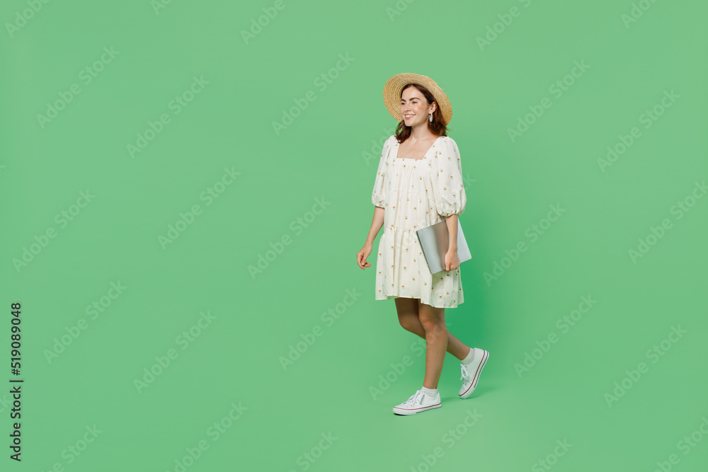 Full body young happy smiling fun woman she 20s wears white dress hat hold use closed laptop pc computer walking going isolated on plain pastel light green background studio. People lifestyle concept. Full body young happy smiling fun woman she 20s wears white dress hat hold use closed laptop pc computer walking going isolated on plain pastel light green background studio. People lifestyle concept.