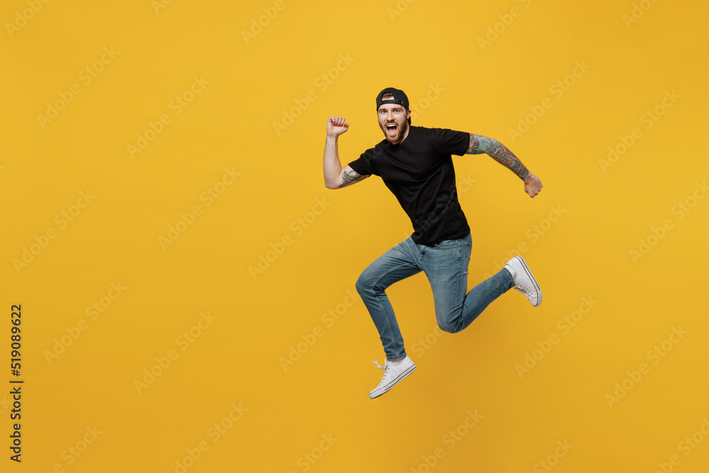 Full body side view sporty strong young bearded tattooed man 20s he wears casual black t-shirt cap jump high run fast isolated on plain yellow wall background studio portrait People lifestyle concept