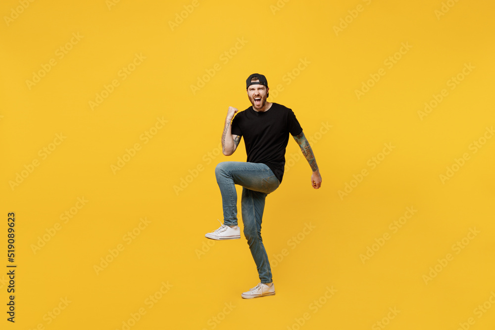 Full body young happy bearded tattooed man 20s he wears casual black t-shirt cap doing winner gesture celebrate clenching fists say yes isolated on plain yellow wall background. Tattoo translate fun.