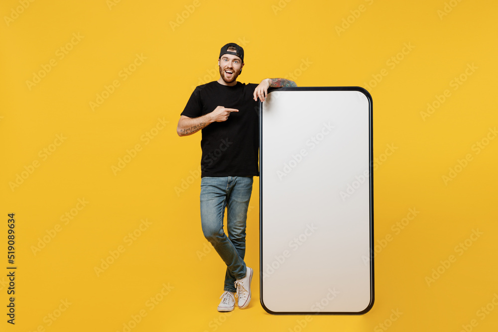 Full body young tattooed man he wears black t-shirt cap point index finger on big huge blank screen mobile cell phone workspace mockup area isolated on plain yellow background Tattoo translate fun - obrazy, fototapety, plakaty 