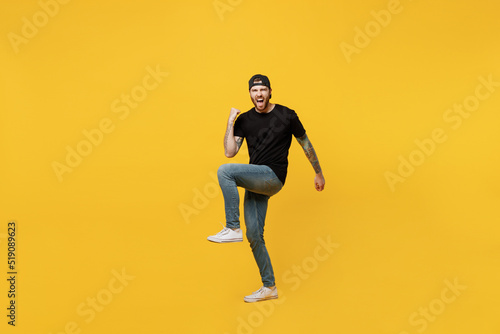 Full body young happy bearded tattooed man 20s he wears casual black t-shirt cap doing winner gesture celebrate clenching fists say yes isolated on plain yellow wall background. Tattoo translate fun.