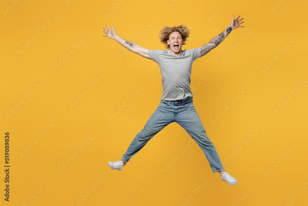 Full body excited exultnt jubilant young caucasian man 20s he wear grey t-shirt look camera jump high with outstretched arms hands isolated on plain yellow backround studio. People lifestyle concept.