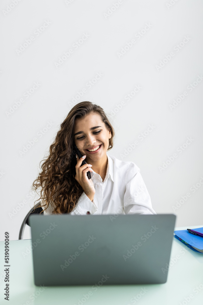 Cheerful young beautiful woman in glasses talking on mobile phone and using laptop with smile while sitting at her working place Cheerful young beautiful woman in glasses talking on mobile phone and using laptop with smile while sitting at her working place