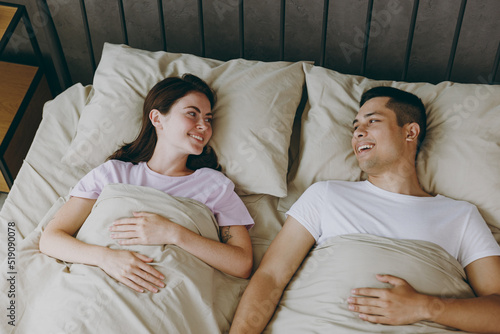 Top view from above happy young couple two family man woman she he wear t-shirt pajama look to each other lying in bed rest spend time together in bedroom home in own room hotel. Real estate concept. © ViDi Studio